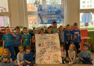 Motylki pozują ubrani na niebiesko na tle dekoracji z okazji Światowego Dnia Świadomości autyzmu trzymając wykonany przez siebie plakat.