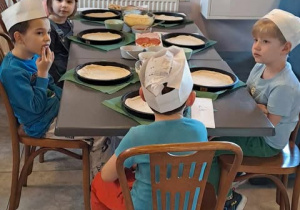 Dzieci siedzą przy stole przed nimi na talerzach leżą ciasta oraz dodatki do pizzy.