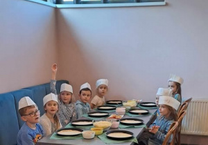 Dzieci siedzą przy stoliku na stole leżą dodatki oraz ciasto na pizzę.