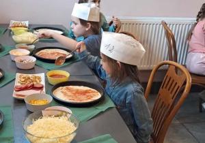 Dzieci siedzą przy stole i smarują ciasto na pizzę sosem pomidorowym.
