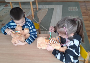 Dziewczynka z chłopcem siedzą przy stoliku i myją misiom zęby.