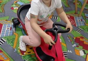 Zdjęcie przedstawia dziewczynkę korzystającą z zabawki jeżdżącej.