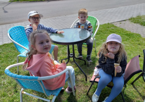 Dzieci zajadają lody przy siedząc przy stoliczkach.