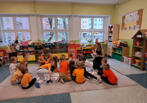 Dzieci siedzą na dywanie w kole na środku leżą dynie oraz kartki z obrazkami dyni.