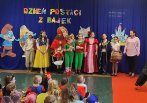 Dziewczynki biorące udział w przedstawieniu oraz ich wychowawczyni stoją na tle dekoracji Dzień Postaci z Bajek.