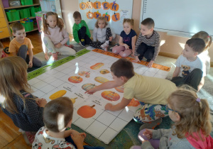 Zdjęcie przedstawia dzieci siedzące na dywanie wokół maty do kodowania na której rozłożone są ilustracje przedstawiające różne odmiany dyni.