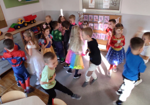 Dzieci przebrane w kolorowe stroje tańczą na dywanie, trzymając w dłoniach połowy ilustracji przedstawiających bajkowe postacie.