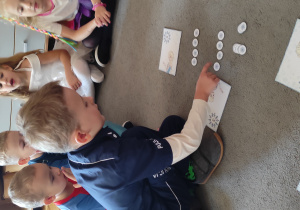 Chłopiec siedzący na dywanie wykonuje działanie matematyczne z wykorzystaniem liczmanów- wyciętych z papieru płatków śniegu. W tle dwóch chłopców i dwie dziewczynki przyglądają się działaniom tego chłopca.