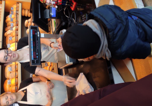 Chłopiec stojący przy ladzie podaje ekspedientce pieniądze za zakupione produkty.