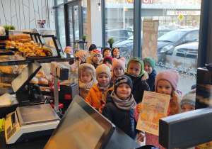 Przedszkolaki ustawione przy ladzie sklepowej pozują do zdjęcia, jedna dziewczynka trzyma w dłoniach podziękowania dla ekspedientki.