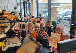 Przedszkolaki ustawione przy ladzie sklepowej pozują do zdjęcia, jedna dziewczynka trzyma w dłoniach podziękowania dla ekspedientki.