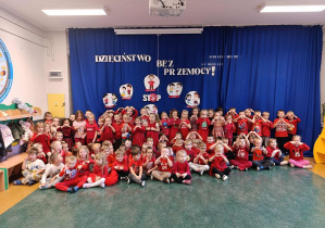 Dzieci z grup trzylatków, czterolatków oraz pięciolatków, ubrane w czerwone koszulki i trzymając ręce ułożone w kształcie serca, pozują do zdjęcia na tle informacji o kampanii Dzieciństwo Bez Przemocy.