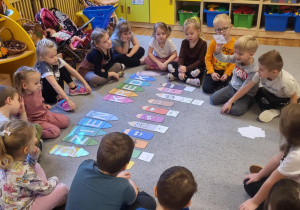 Dzieci siedzą na dywanie, chłopiec nazywa co jest na rysunki i dopasowuje do kredki w odpowiednim kolorze.