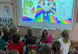 Dzieci oglądają na tablicy multimedialnej film edukacyjny dotyczący kredek i kolorów.