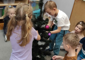 Troje przedszkolaków stojących wkoło choinki wiesza na niej ozdoby.