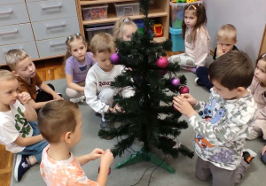 Troje przedszkolaków stojących wkoło choinki wiesza na niej ozdoby.