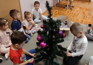 Dwaj chłopcy siedzący wkoło choinki wiesza na niej ozdoby. Pozostałe dzieci siedzą na dywanie i przyglądają się.