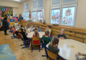 Zdjęcie przedstawia dzieci siedzące przy stoliczkach w sali przedszkolnej, przedszkolaki uważnie słuchają Pani prowadzącej warsztaty, która tłumaczy jak wykonać stroik.