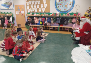 Zdjęcie przedstawia dzieci siedzące na dywanie przed Św. Mikołajem i czekające na swoje upominki