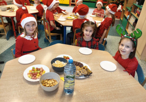 Dzieci siedzą przy stolikach, a na stołach czeka na nie przygotowany poczęstunek.