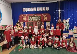 Dzieci pozują do zdjęcia grupowego na tle świątecznej dekoracji wraz z panią oraz Świętym Mikołajem, trzymając w rękach prezenty.