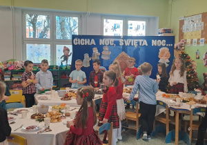 Dzieci stoją przed stolikami na stołach leżą potrawy wigilijne.