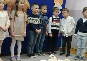 Chłopcy w odświętnych strojach oraz dziewczynki w strojach aniołków stoją na tle dekoracji świątecznej. Przed nimi stoi żłóbek.