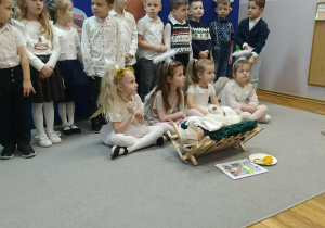 Dziewczynki w strojach aniołków siedzą przed żłóbkiem. Za nimi przedszkolaki stoją na tle świątecznej dekoracji.