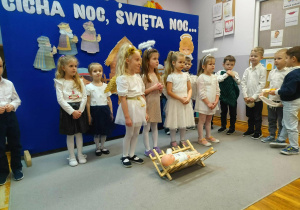 Cztery dziewczynki w strojach aniołków stoją przed żłóbkiem. Za nimi dzieci w odświętnych strojach stoją na tle bożonarodzeniowej dekoracji.