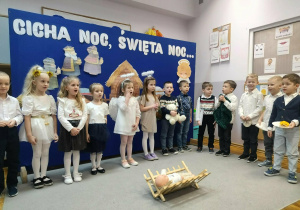 Uśmiechnięte dzieci stojące wokół żłóbka śpiewają kolędę.