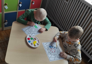 Dwóch chłopców siedzi przy stoliku, malują farbami obrazek ze śnieżynką