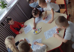 Dzieci siedzące przy stolikach wykonują pracę plastyczną z wykorzystaniem farby.