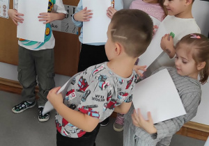 Dzieci stoją na dywanie i próbują utrzymać białe kartki na brzuchu.