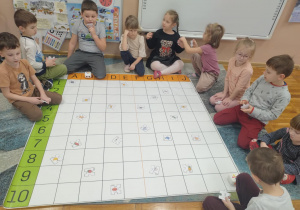 Zdjęcie przedstawia dzieci siedzące na dywanie wokół maty do kodowania. Na macie rozłożone są puzzle z obrazkami.