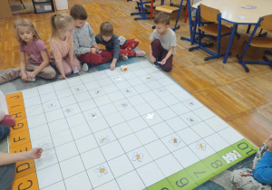 Zdjęcie przedstawia dzieci siedzące przed matą gdzie rozłożone są obrazki.