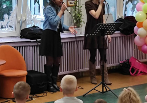 Dwie młode kobiety śpiewają dla dzieci trzymając mikrofony w dłoniach.