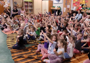 Dzieci siedzące na kocach machają uniesionymi rękoma.