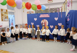 Przedszkolaki stoją w półkolu i recytują wiersz dla babci i dziadka.