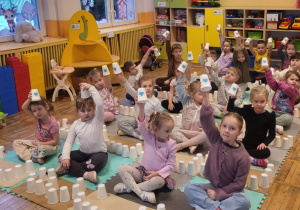 Dzieci siedzą na dywanie, trzymają w górze kubeczek z niebieskim serduszkiem.