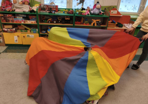 Kolorowa chusta przykrywa bawiące się dzieci w sali przedszkolnej.
