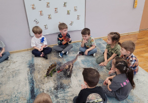 Słoneczka siedzą na dywanie i witają się z dinozaurem