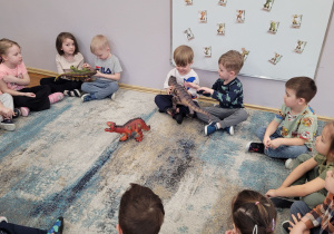 Słoneczka siedzą na dywanie i oglądają figurki dinozaurów.