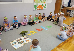 Dzieci siedzą w kola pośrodku którego lezą różnokolorowe dinozaury z jajami posegregowanymi ze względu na kolor.