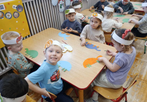 Dzieci siedzą przy stoliczku i doklejają elementy papierowego dinozaura.