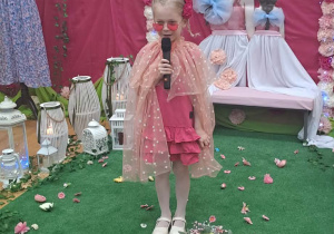 Dziewczynka trzyma w ręku mikrofon stoi w tle dekoracji w pięknym stroju.