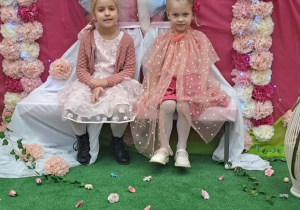 Dziewczynki siedzą na ławeczce, pozują do zdjęcia w tle dekoracji.