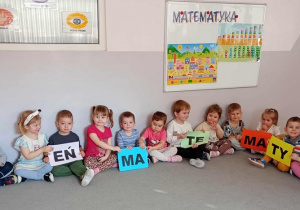 Przedszkolaki siedzą w półkolu na dywanie i trzymają w rękach napis dzień matematyki.