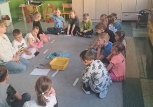 Dzieci siedzą razem za panią w kole w środku leżą przedmioty.