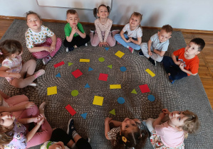 Dzieci siedzą w kole. W środku kolorowe figury geometryczne