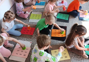 Dzieci siedzą przed dywanikami i bawią się w matematyczną zabawę z kubeczkami.